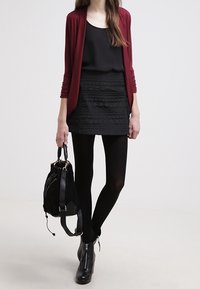 Červený cardigan, černý top bez rukávů, texturovaná černá mini sukně, černé punčocháče, kotníkové boty a černý batoh. Jednoduchý, vrstvený outfit.