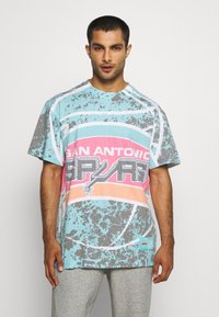 Mand iført en løs pastelblå og pink San Antonio Spurs grafisk T-shirt og lysegrå sweatpants, som står med ansigtet mod fronten med neutral udtryk.