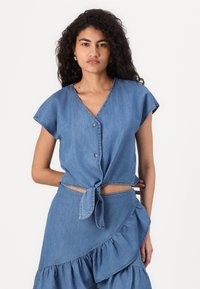 Blå denim kortermet topp med V-hals, knapplukking og knyte-detalj ved livet, kombinert med et matchende volangskjørt.