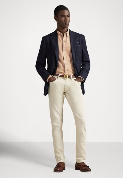 Vīrietis, kurš valkā tumši zilu žaketi, gaiši brūnu pogājamu kreklu, bēša krāsas bikses, brūnu jostu un brūnas kurpes ar pušķiem, stāv ar rokām kabatās.