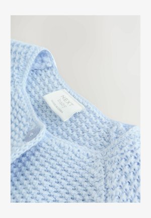Világoskék, kötött kisbaba kardigán gombbal és "Next baby" címkével a nyakában, fehér háttéren bemutatva.