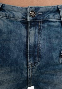 Jeans in denim blu con chiusura a bottone, tasche frontali e cuciture a contrasto. Il tessuto presenta un aspetto leggermente usurato con tonalità variabili.