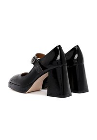 Chaussures à talons carrés en cuir verni noir avec un bout arrondi et une sangle réglable ornée d'une boucle métallique circulaire. Fini lisse et brillant.