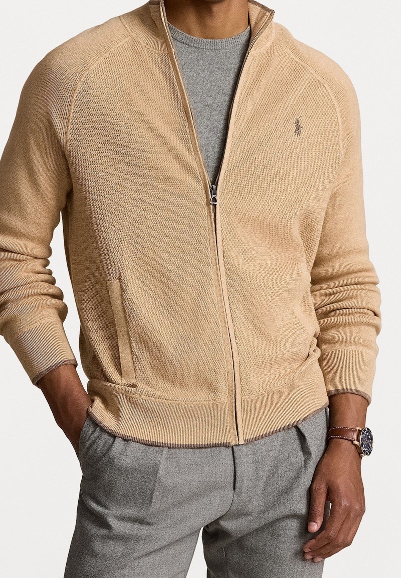 Homme portant un pull en tricot beige zippé sur une chemise grise, un pantalon gris et une montre en cuir marron au poignet gauche, les mains dans les poches.