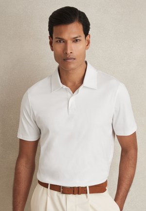 Bărbat cu păr scurt și întunecat poartă un tricou polo alb cu mâneci scurte băgat în pantaloni bej cu o curea maro din piele, stând în fața unui fundal neutru.