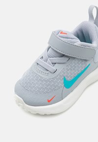 Sivi atletski tenisica s mrežastim gornjim dijelom, tirkiznim Nike swoosh-om, crvenim detaljima i bijelim potplatom. Ima čičak trakicu i dizajn s vezicama.