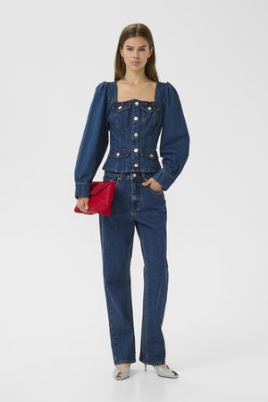 Mujer con chaqueta abotonada de denim azul oscuro con mangas abullonadas y jeans a juego, sosteniendo un clutch rojo y llevando tacones gris claro.