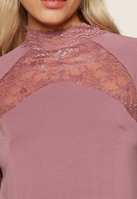 Haut lilas avec un col en dentelle haute et un motif floral, en tissu lisse, avec une silhouette ajustée, mettant en valeur la texture et les détails de conception.