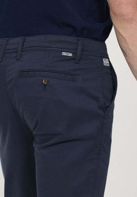 Pantalons en coton bleu marine à coupe droite, avec un bouton marron et des coutures décoratives. Comprend des poches latérales et un détail de poche arrière.