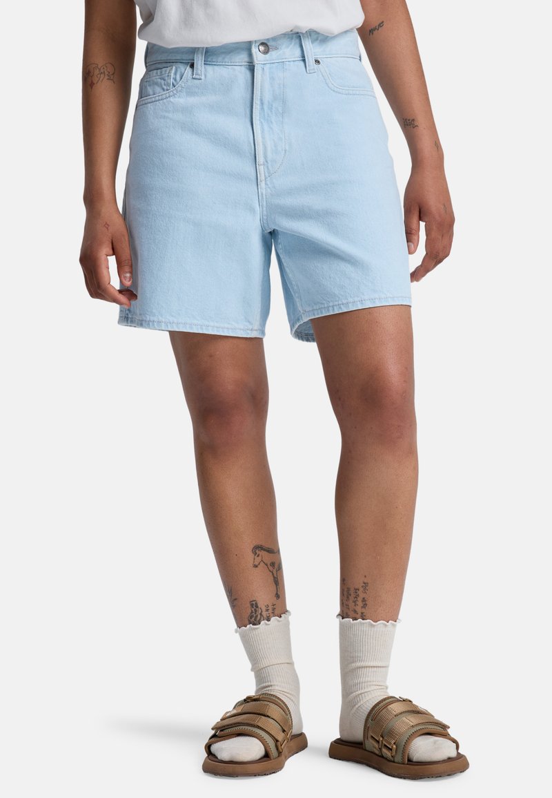 Ανοιχτό μπλε τζιν σορτς με κανονική εφαρμογή, ψηλή μέση, απλή ραφή και δύο μπροστινές τσέπες. Συνδυασμένο με καφέ σανδάλια και λευκές κάλτσες.