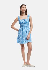 Blauwe bloemenjurk met een vierkante halslijn, opgeblazen bandjes, elastische taille en een uitlopende rok, gemaakt van lichte stof. Platte beige schoenen.