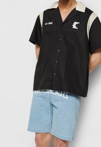 Czarna koszula z krótkim rękawem zapinana na guziki z beżowym kołnierzykiem; posiada białą haftowaną aplikację "est. 1989" oraz logo, w zestawie z jasnoniebieskimi szortami dżinsowymi.