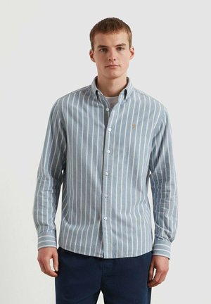 Giovane uomo che indossa una camicia button-down a righe verticali azzurro chiaro e bianca con maniche lunghe e pantaloni blu scuro, in piedi su uno sfondo semplice.