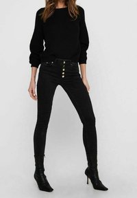 Schwarzer, gerippter Pullover mit rundem Halsausschnitt, kombiniert mit hochgeschnittenen, engen schwarzen Jeans mit fünf Knöpfen vorne und knöchellanger Länge.