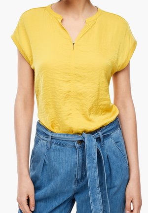 Vrouw draagt een gele blouse met korte mouwen, ingestopt in een hooggetailleerde blauwe spijkerbroek met een gebonden stoffen riem.