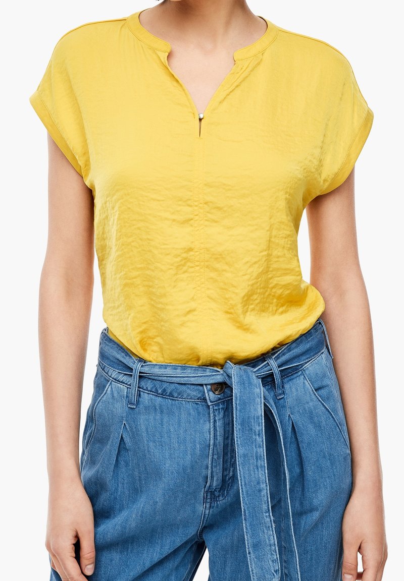 Femme portant un chemisier jaune à manches courtes rentré dans un pantalon en jean bleu taille haute avec une ceinture en tissu nouée.