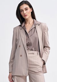 Beżowa marynarka w prążki z podwójnymi guzikami, w połączeniu z dopasowaną satynową bluzką z wysokim kołnierzem i eleganckimi spodniami, tworząc skoordynowaną całość.