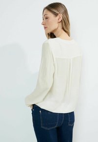 Kermainen pitkähiha blouse, jossa on tekstuurinen kangas, pyöreä pääntie ja takana laskosyksityiskohta, yhdistettynä tummansinihin farkkuihin.