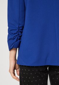 Modrý top z texturované, pružné látky; má nařasené rukávy. Nošeno s černými kalhotami zdobenými hvězdnými vzory.