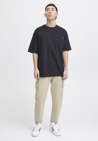 Zwarte oversized katoenen t-shirt met een borstzak, gecombineerd met lichtbeige cargobroek met zijzakken en boorden, gedragen met witte sneakers.