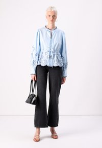 Blusa azzurro chiaro con ricami floreali e vita arricciata, abbinata a jeans svasati scuri e una piccola borsa nera. Tessuto con una texture liscia.