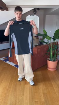 Persoană tânără îmbrăcată cu pantaloni bej și adidași, ținând o cămașă cu mânecă lungă neagră și gri într-un living modern, cu o plantă și o canapea.