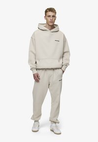 Beige hettegenser med frontlomme og svart logo, kombinert med matchende beige joggebukser. Modellen har på seg hvite joggesko. Stoffet ser mykt og teksturert ut.