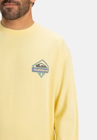 Yellow long-sleeve sweatshirt with a crew neck, featuring a blue and turquoise Quiksilver logo on the chest. Soft fabric, relaxed fit.