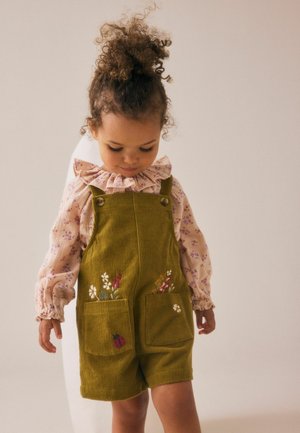 Grüne Cord-Dungarees mit zwei Vordertaschen, verziert mit floraler Stickerei und einem Marienkäfer-Detail. Kombiniert mit einer Bluse im hellen Blumenmuster.