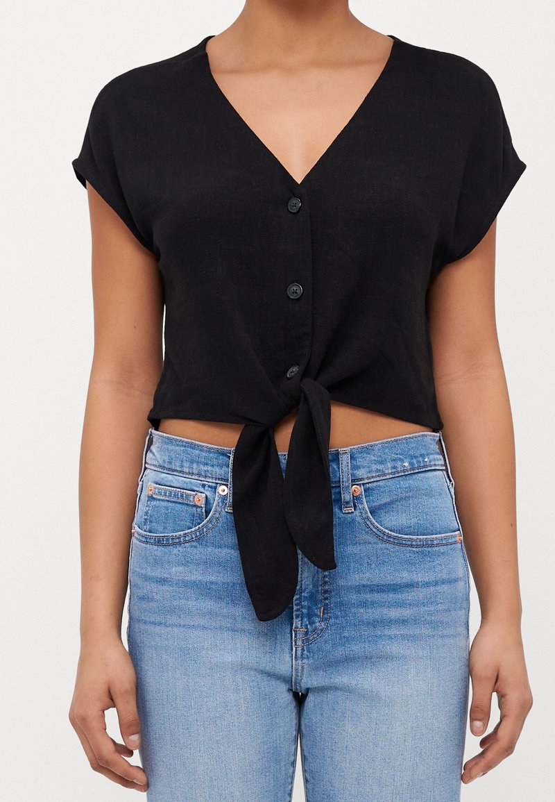 Donna che indossa un top corto nero con maniche corte e bottoni, annodato, abbinato a jeans a vita alta di un blu chiaro.