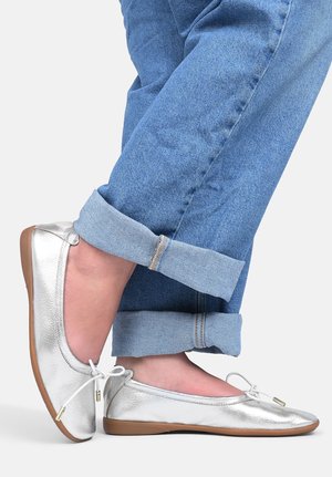Stopy w srebrnych balerinach z białymi sznurowadłami i podwiniętymi niebieskimi dżinsami na prostym tle.