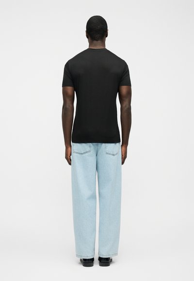Άνδρας που φοράει απλό μαύρο κοντομάνικο μπλουζάκι, ανοιχτό μπλε φαρδιά τζιν παντελόνια και μαύρα παπούτσια, στέκεται με την πλάτη σε λευκό φόντο.