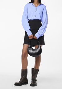 Nainen, jolla on päällään sininen paita, musta minihame, jossa on sivureikä, tummanruskeat solkilngät ja joka pitää käsissään mustaa puolikuun muotoista käsilaukkua.