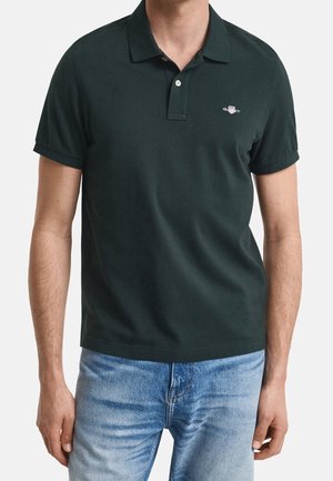 Mężczyzna noszący ciemnozieloną koszulkę polo z krótkim rękawem i małym logo na piersi oraz jasnoniebieskie jeansy, stojący na jednolitym tle.