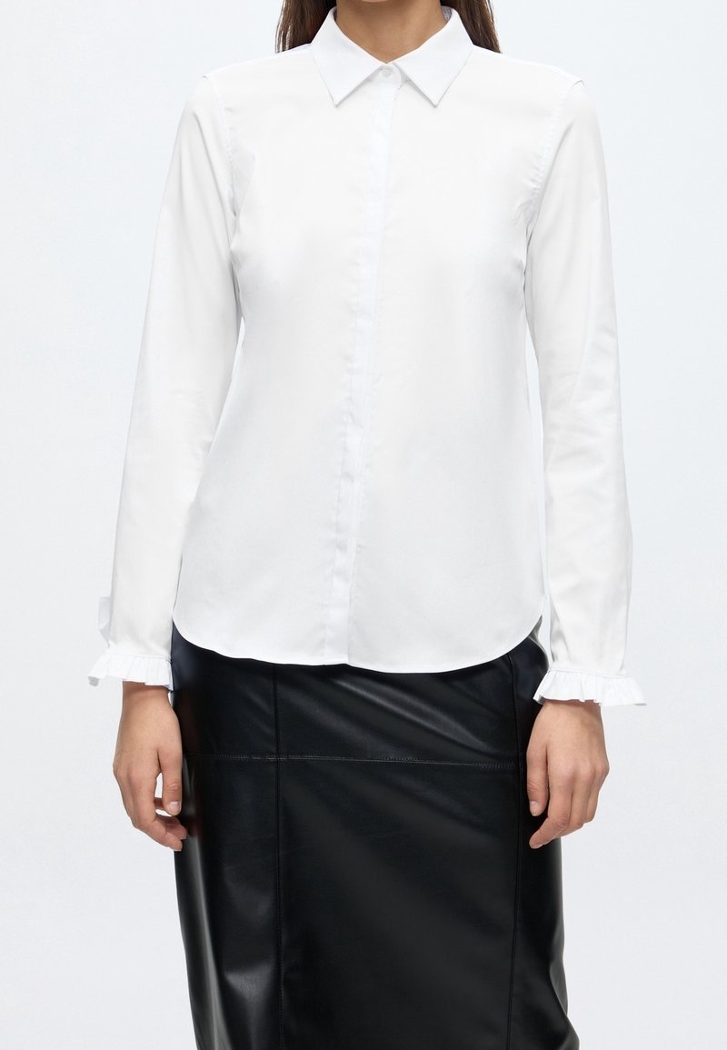 Femme portant un chemisier blanc à manches longues boutonné avec des poignets à volants et une jupe en cuir noire longueur genou.