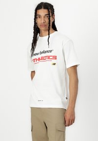 Baltas īsām piedurknēm T-kreklī, uz kura ir melni un sarkani uzraksti "new balance" un "ATHLETICS" ar dzeltenu logo akcentu. Vienkāršs, ikdienišķs dizains.