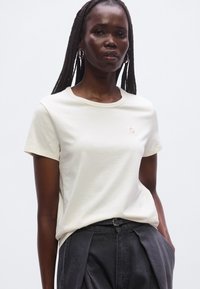 Kremowy t-shirt z krótkim rękawem wykonany z miękkiego materiału, z okrągłym dekoltem i małym logo na piersi.