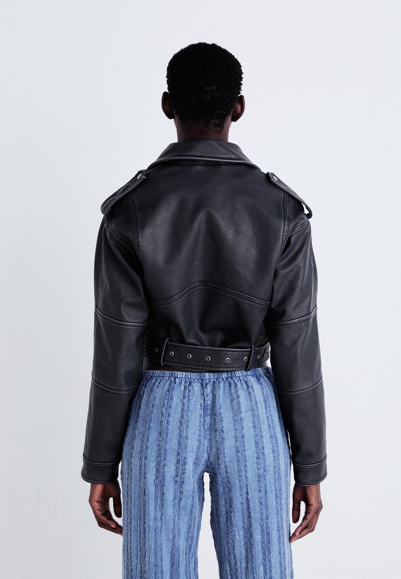 Veste cropped en similicuir noir avec épaulettes et taille ceinturée. Portée avec un pantalon large à texture bleue. Vue arrière.