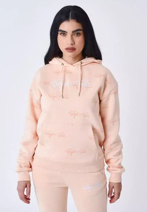 Vrouw met een lichtperzikkleurige hoodie en bijpassende joggingbroek met "Project x Paris" logo's, staand tegen een effen witte achtergrond.