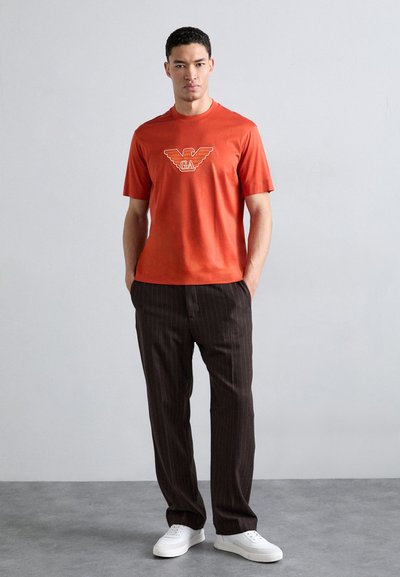 Oranžinė medvilninė marškinėliai su trumpomis rankovėmis ir logotipo grafika ant krūtinės, derinami su rudomis juostelėmis kelnių ir baltais sportbačiais.