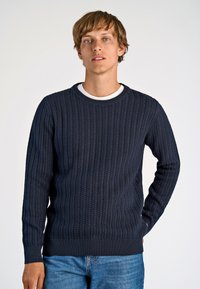 Marineblauer gestrickter Pullover mit einem strukturierten Muster und gerippten Bündchen und Saum. Getragen über einem weißen Rundhalsausschnitt-Shirt, kombiniert mit blauen Jeans.