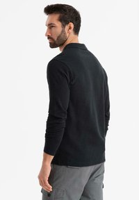 Černá polo košile s dlouhými rukávy a límečkem, obepnutý střih. Měkká textura a volný střih; viditelné švy a minimalistické stylové detaily.