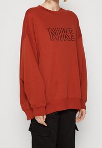 Person som bär en överdimensionerad bränd orange Nike-tröja med svarta cargobyxor, avskuren vid nacken och mitt på låret framför en neutral bakgrund.