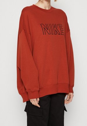 Person som bär en överdimensionerad bränd orange Nike-tröja med svarta cargobyxor, avskuren vid nacken och mitt på låret framför en neutral bakgrund.