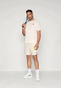 Šviesiai smėlinio spalvos sportinis marškinėlių ir šortų komplektas su mėlynu logotipu ant krūtinės. Balti kojinės ir batai, laikantis teniso raketę ir kamuoliuką.