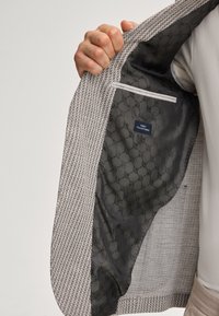 Leicht strukturierter beiger Blazer mit einem gemusterten, dunkelgrauen Futter. Die Innenseite verfügt über eine Tasche und ein Etikett mit der Aufschrift "Finest Italian Fabric."