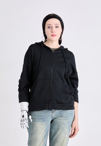 Crna zip-up dukserica s prednjim džepom, kapuljačom s vezicom i opuštenim krojem, uparena s prugastim svijetlim farmerkama i robotskom rukom.