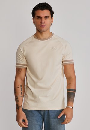 Mann mit kurzen dunklen Haaren, der ein beigefarbenes T-Shirt mit kurzen Ärmeln und gestreiften Bündchen sowie blaue Jeans trägt, steht vor einem schlichten hellen Hintergrund.