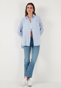 Camicia azzurro chiaro a maniche lunghe con bottone e tasca, abbinata a jeans svasati in lavaggio chiaro e sneakers bianche. Tessuto liscio e strutturato.
