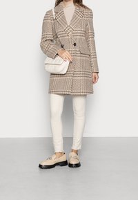 Cappotto doppiopetto a quadri beige, dolcevita crema, pantaloni bianchi slim-fit e mocassini beige con suola nera, mentre porta una borsa a tracolla trapuntata crema.